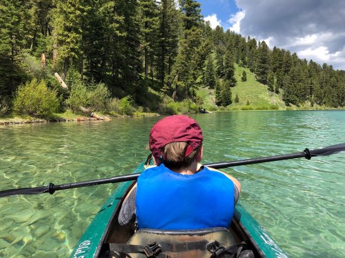 A person in a kayak paddles on a clear lake surrounded by lush green trees and hills under a partly cloudy sky.
