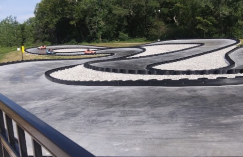 A winding go-kart track with two karts racing on a sunny day, surrounded by greenery.