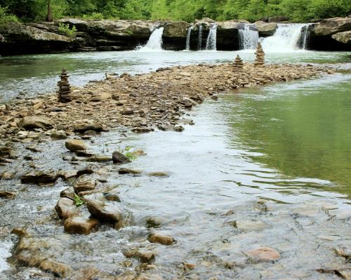 A serene river scene with stacked stones on a rocky shore and two small waterfalls in the background, surrounded by greenery.