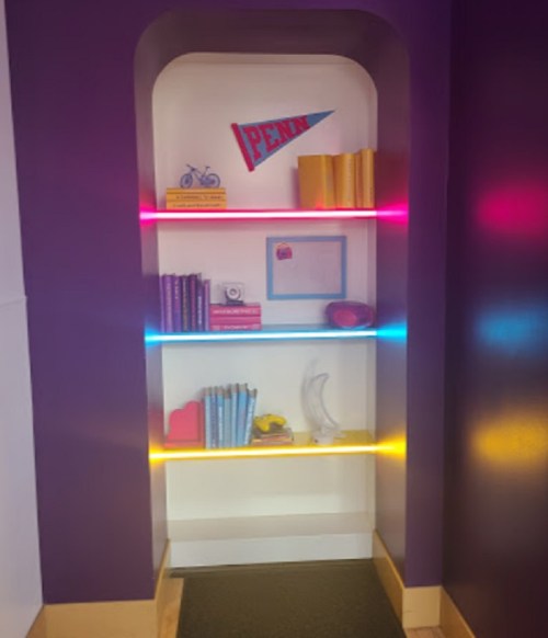 A colorful bookshelf with illuminated shelves, featuring books, a pennant, and decorative items against a purple wall.