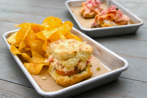 A biscuit sandwich filled with lobster and tomato, served with crispy potato chips on a metal tray.