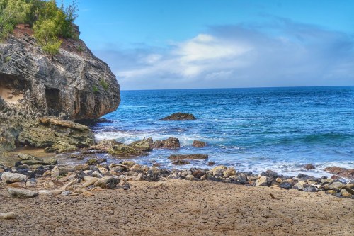 A rocky beach with gentle waves, cliffs on one side, and a clear blue sky above.