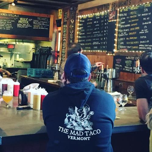 A person in a hoodie sits at a bar, looking at a menu board with various drink options and specials.