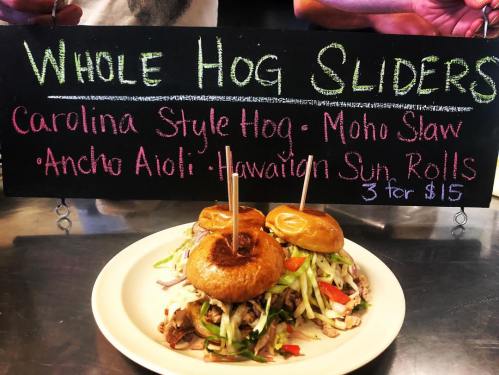 A plate of three sliders with pulled pork, slaw, and sauces, displayed in front of a colorful chalkboard menu.