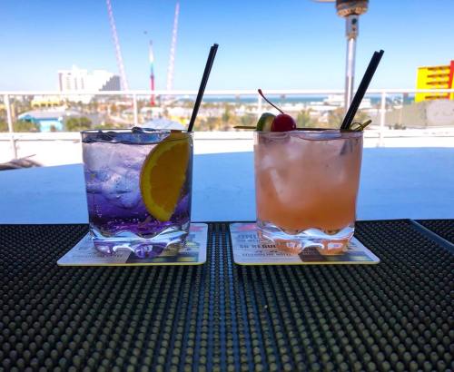Two colorful cocktails on a table with a beach view in the background, one with a lemon slice and the other with cherries.