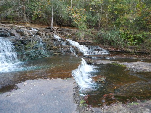 A serene waterfall cascades over rocky ledges into a calm pool, surrounded by lush greenery and autumn leaves.