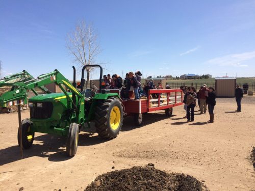 A green tractor with a red wagon is parked on dirt, while a group of people gathers around in a sunny outdoor setting.