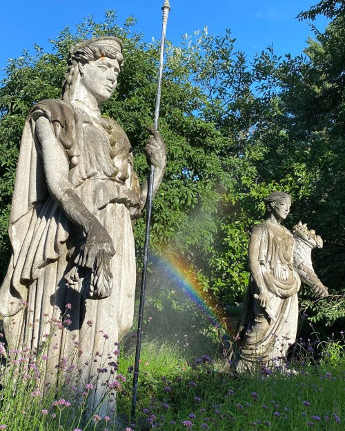 Two weathered statues stand in a garden, surrounded by greenery and flowers, with a rainbow arcing in the background.