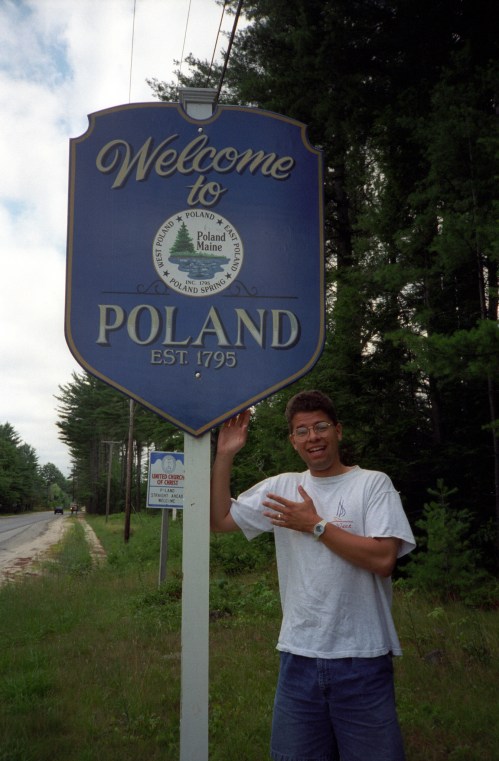 A person stands next to a "Welcome to Poland" sign in a wooded area, smiling and gesturing towards the sign.