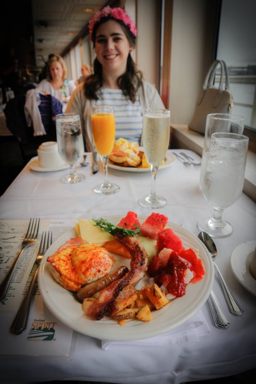 A table set for brunch with a plate of food, drinks, and a smiling woman in the background wearing a flower crown.