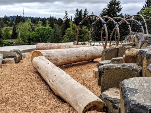 Playground area featuring large logs, rocks, and climbing structures, surrounded by trees under a cloudy sky.