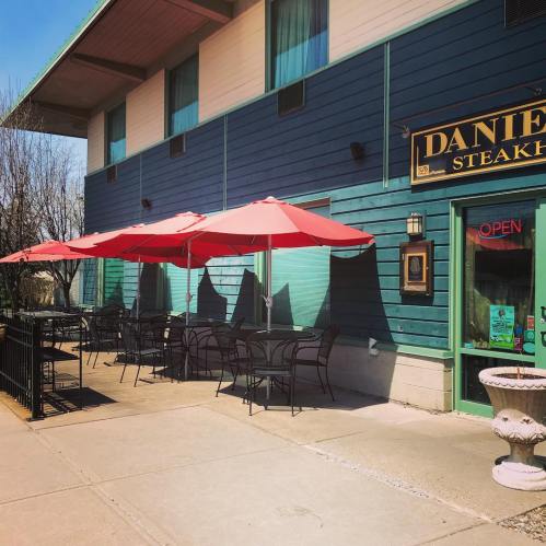 Outdoor dining area of a steakhouse with red umbrellas and black chairs, set against a blue wooden building.