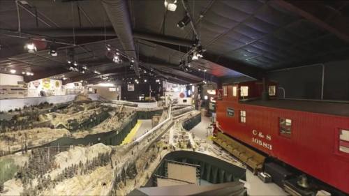 A panoramic view of a model train exhibit featuring detailed landscapes and a red train car in a large indoor space.