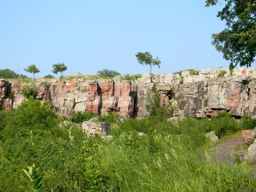 A rocky cliff with patches of greenery and small trees against a clear blue sky.