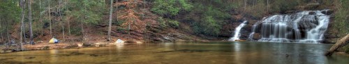 A serene campsite by a waterfall, surrounded by lush trees and a calm river, with tents set up nearby.