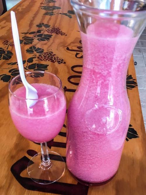 A glass and a pitcher filled with a pink, frothy beverage on a wooden table. A white spoon rests in the glass.