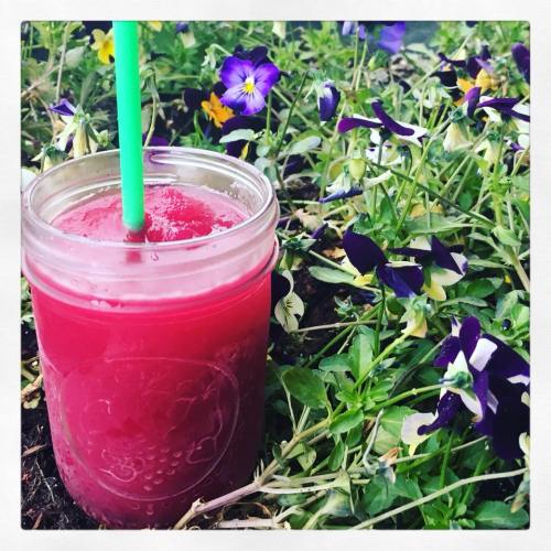 A vibrant pink smoothie in a jar with a green straw, surrounded by purple and green flowers.