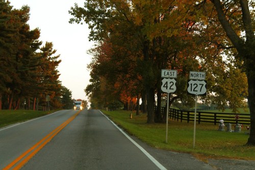 A rural road with signs for East 42 and North 127, lined with trees and a glimpse of a truck in the distance.