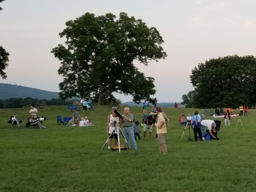 A group of people set up telescopes on a grassy field, with trees and hills in the background during twilight.