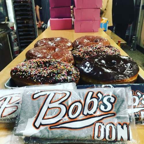 A tray of colorful donuts with sprinkles and chocolate icing, alongside Bob's Donuts merchandise on a table.
