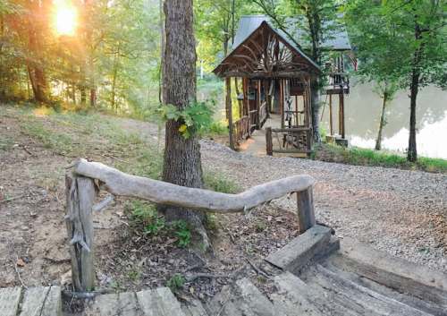 A rustic wooden railing leads down steps to a serene riverbank, with a cabin nestled among trees in the background.