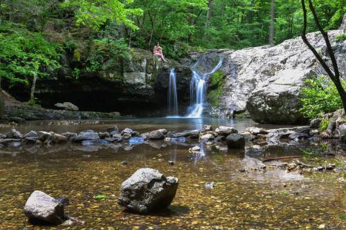A serene waterfall cascades into a clear pool surrounded by lush greenery and rocky terrain. A person sits nearby.