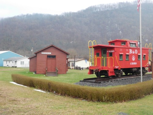 A red caboose and a small wooden building on a grassy area, with mountains in the background and a flagpole nearby.