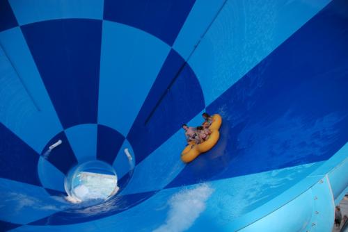 Two people on a yellow inner tube slide down a blue water slide, surrounded by a spiral pattern.
