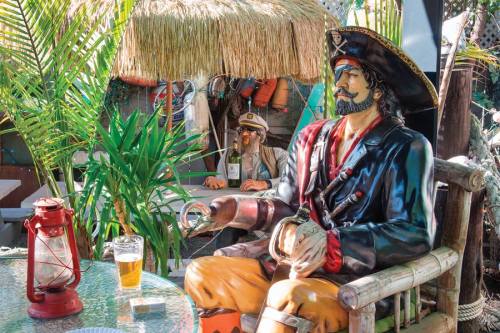 A pirate statue sits in a tropical setting with a drink, while another figure in a captain's hat is in the background.