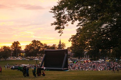 A large inflatable screen set up for an outdoor event at sunset, with a crowd gathered on the grass.