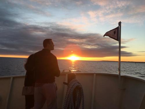A couple stands on a boat, watching a vibrant sunset over the water, with clouds and a flag in the foreground.