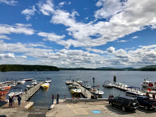 A scenic view of a lake with boats docked, under a partly cloudy sky and surrounded by green hills.