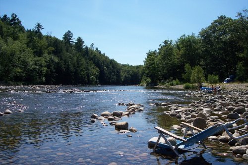 A serene river scene with smooth stones, lush trees, and a blue chair by the water under a clear blue sky.