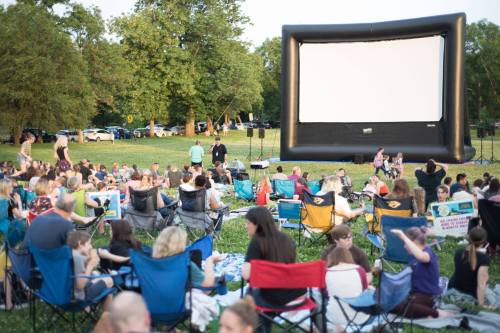 A large outdoor gathering in a park with people sitting on blankets and chairs, facing a large inflatable movie screen.