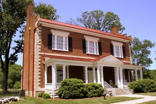 Historic two-story brick house with a red metal roof, white columns, and lush green landscaping.
