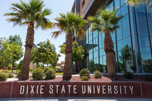 Sign for Dixie State University in front of a modern building, surrounded by palm trees and greenery.