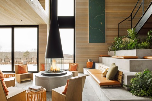 Modern interior with a central fireplace, seating areas, large windows, and greenery, creating a cozy atmosphere.