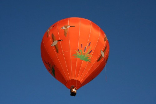 A vibrant orange hot air balloon with bird designs floats against a clear blue sky.