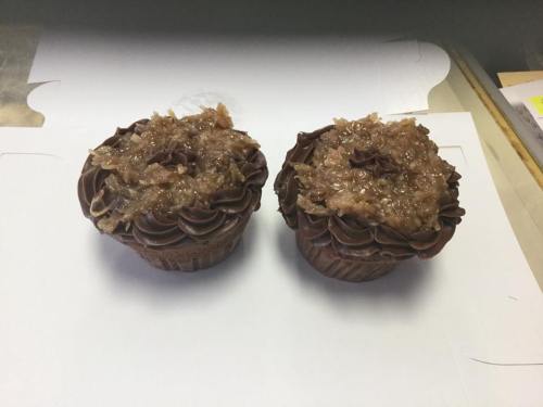 Two chocolate cupcakes topped with a glossy coconut and pecan frosting, placed on a white surface.