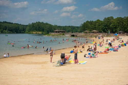 A sandy beach with people sunbathing, swimming, and relaxing by a calm lake surrounded by trees and pavilions.