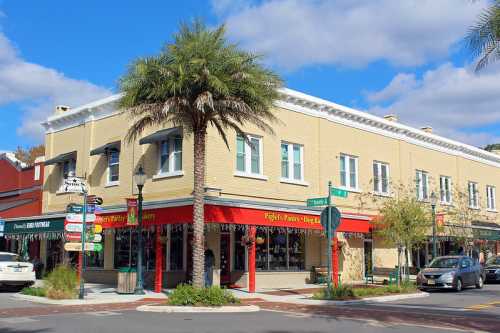 A corner building with a red awning, palm tree, and street signs in a sunny, vibrant downtown area.