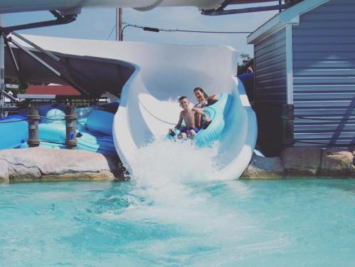 Two boys slide down a water slide, splashing into a bright blue pool on a sunny day.