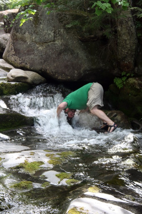 A person in a green shirt bends over a stream, playfully interacting with the water near large rocks and greenery.