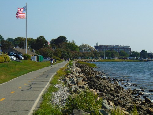 A scenic waterfront path with a flag, cyclists, and a view of a building along the shoreline.