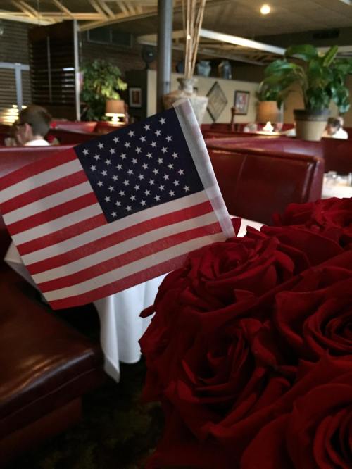 A small American flag stands among a bouquet of red roses in a restaurant setting.