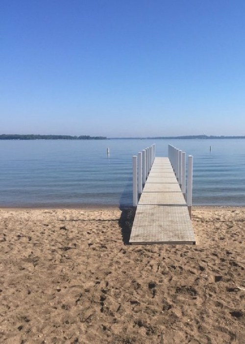 A wooden dock extends over calm water, leading to a clear blue sky and sandy beach.