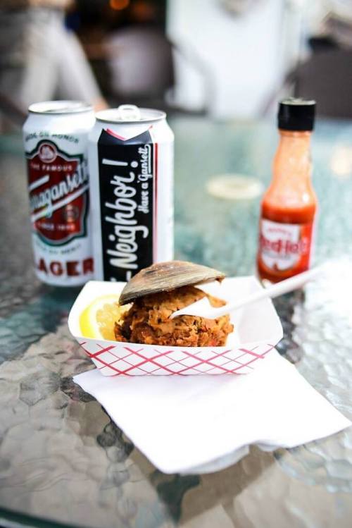 A seafood dish with a shell on top, served with a lemon wedge, hot sauce, and two cans of beer on a glass table.