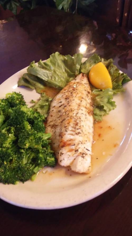 A plate featuring a seasoned fish fillet on lettuce, accompanied by steamed broccoli and a lemon wedge.