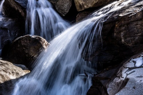 A close-up of a waterfall cascading over dark rocks, with sunlight illuminating the flowing water.