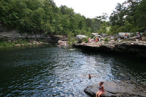 A serene swimming area surrounded by rocky cliffs and lush greenery, with people enjoying the water and sunbathing.